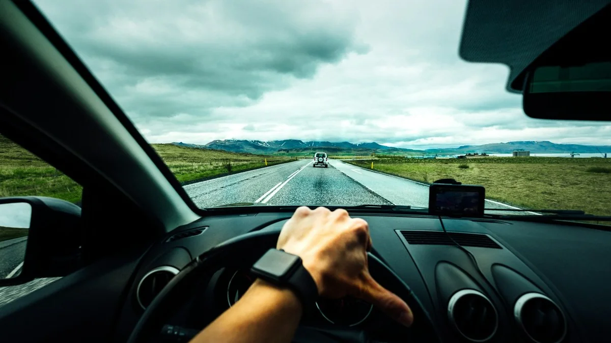 The view from a driver's seat: the road and a car ahead through the windscreen, the dashboard below it, and both hands on the steering wheel in the foreground